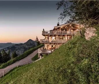 Ein gemütliches Chalet auf einem Hügel mit vielen Balkonen. Im Hintergrund erstreckt sich eine malerische Berglandschaft bei Sonnenuntergang.