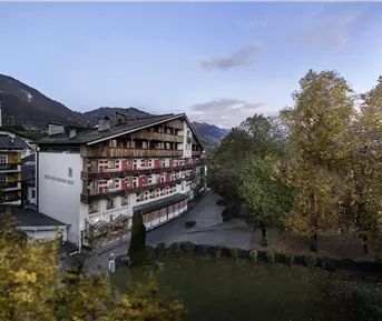 Ein malerisches Hotelgebäude umgeben von Bäumen und majestätischen Bergen. Der Himmel ist klar und die Atmosphäre ist ruhig und einladend.