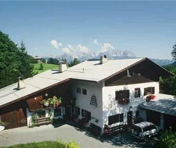 Ein gemütliches Holzhaus umgeben von grünen Bäumen und Bergen im Hintergrund. Der Himmel ist blau und klar.